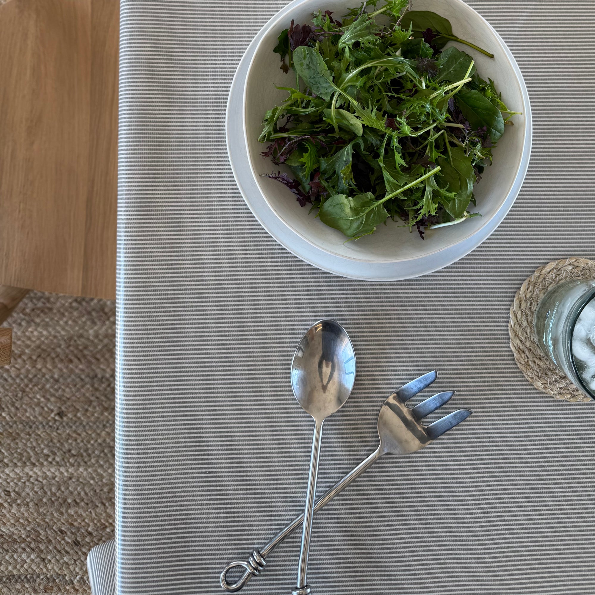 A top-down view of a dining table setting featuring a white bowl filled with green salad, a pair of silver metal serving utensils (spoon and fork) with decorative handles, and a glass on a woven coaster, all arranged on a gray and white striped tablecloth.