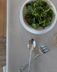 A top-down view of a dining table setting featuring a white bowl filled with green salad, a pair of silver metal serving utensils (spoon and fork) with decorative handles, and a glass on a woven coaster, all arranged on a gray and white striped tablecloth.