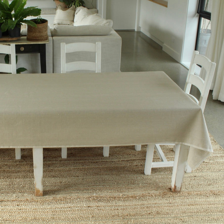 Beige tablecloth on a dining table with white chairs in a room with a rug and plants.