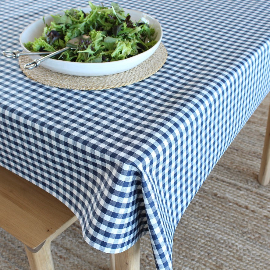 A blue and white gingham checkered tablecloth draped over a wooden dining table, with a white bowl of salad on a woven placemat in the foreground.