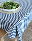 A blue and white gingham checkered tablecloth draped over a wooden dining table, with a white bowl of salad on a woven placemat in the foreground.