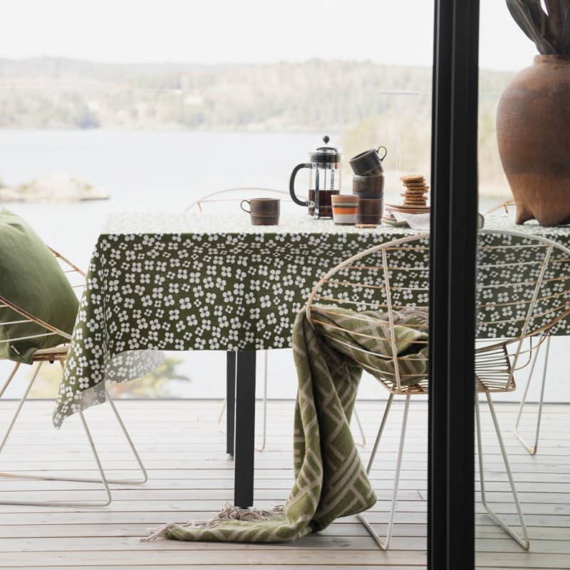 An olive green tablecloth with a white floral pattern is draped over a dining table on a wooden deck, with a matching green throw blanket hanging from the chair. A black French press and mugs sit on the table, with a scenic lake and forest in the background.