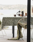 An olive green tablecloth with a white floral pattern is draped over a dining table on a wooden deck, with a matching green throw blanket hanging from the chair. A black French press and mugs sit on the table, with a scenic lake and forest in the background.