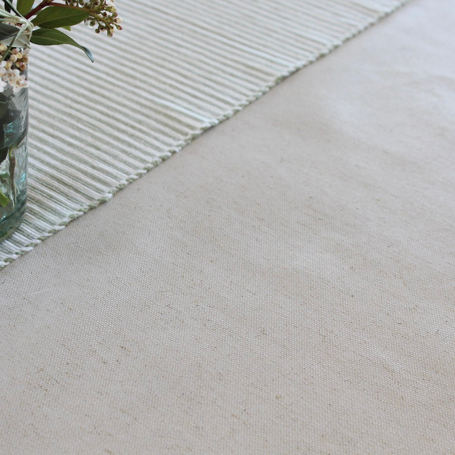 A close-up of a light beige woven textile surface with a striped cream runner or placemat and a small glass vase with flowers in the upper left corner.