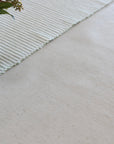 A close-up of a light beige woven textile surface with a striped cream runner or placemat and a small glass vase with flowers in the upper left corner.