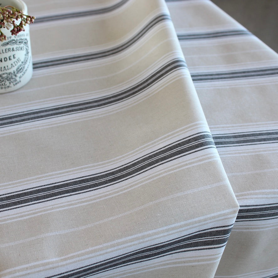 A close-up of a striped tablecloth in cream with thin black and gray horizontal stripes, draped over a table with a small decorative cup visible in the upper left corner.