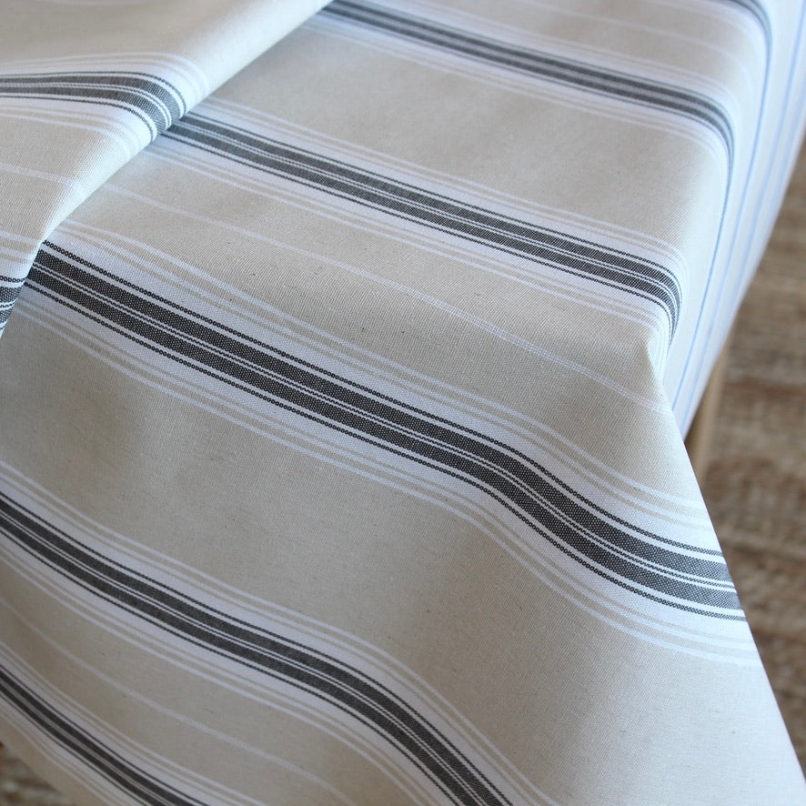 A close-up of a striped tablecloth featuring horizontal stripes in dark gray, black, and white on a warm off-white background.