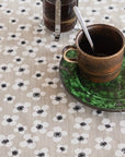 A close-up of a beige tablecloth with a repeating black and white floral pattern, styled with a rustic bronze mug on a green saucer and a dark glass container.