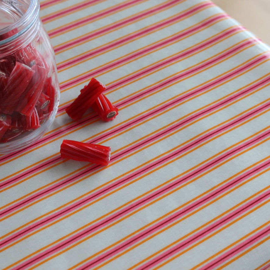 Red licorice candy pieces, some inside a clear glass jar and a few loose, arranged on a white tablecloth with diagonal pink and orange stripes.