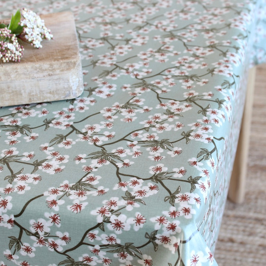 A tablecloth with a delicate floral print of white blossoms on dark branches over a muted sage green background, styled on a table with a rustic wooden block and white flowers.