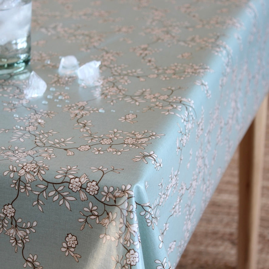 A light blue/green tablecloth with a delicate beige and brown cherry blossom branch pattern, draped over a table with a glass and ice cubes