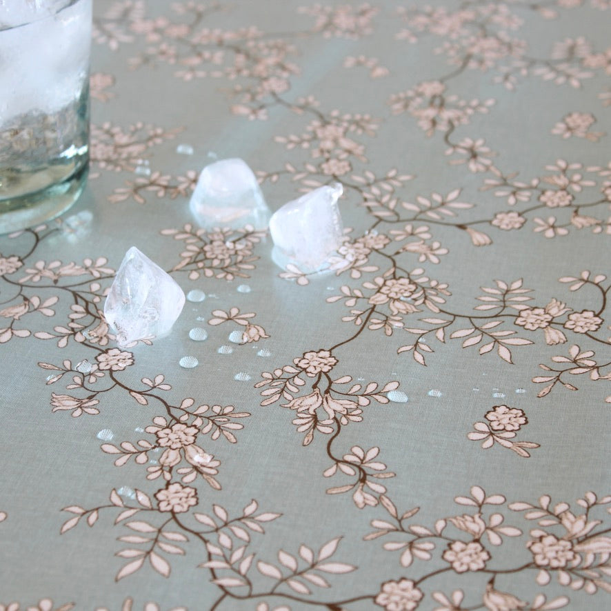 A close-up of a tablecloth featuring a delicate brown and beige floral branch pattern on a pale gray background, with ice cubes resting on the surface.