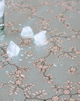 A close-up of a tablecloth featuring a delicate brown and beige floral branch pattern on a pale gray background, with ice cubes resting on the surface.