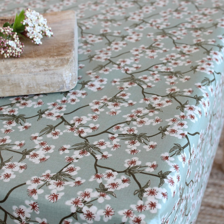 A tablecloth with a delicate cherry blossom branch pattern in white, dusty pink, and sage green on a muted green background, styled with a rustic wooden block and white flowers in the corner.