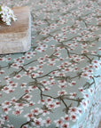 A tablecloth with a delicate cherry blossom branch pattern in white, dusty pink, and sage green on a muted green background, styled with a rustic wooden block and white flowers in the corner.