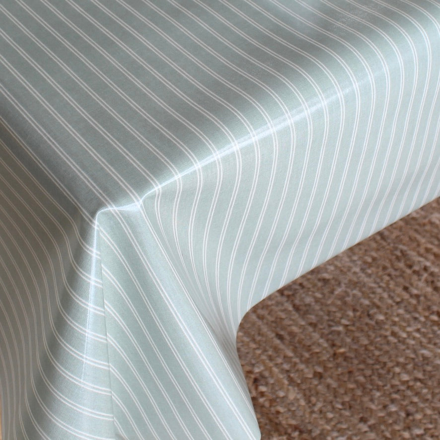 A close-up of a tablecloth with a pale sage green background and thin white vertical pinstripes, draped over the corner of a table with a brown textured carpet visible underneath.