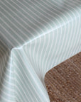 A close-up of a tablecloth with a pale sage green background and thin white vertical pinstripes, draped over the corner of a table with a brown textured carpet visible underneath.