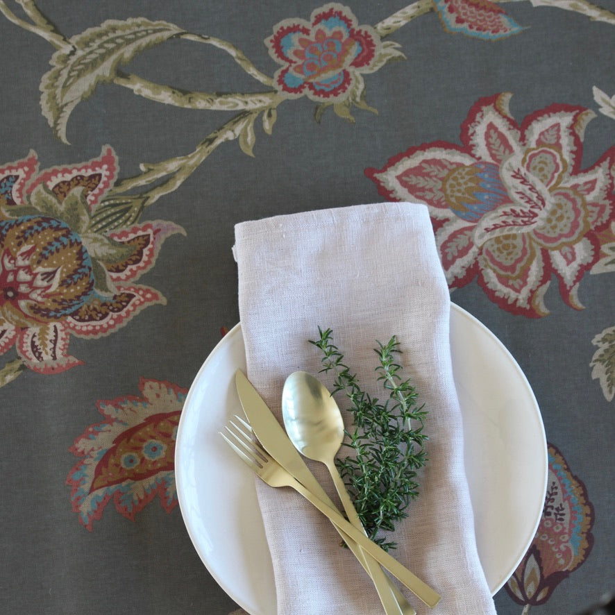 Table setting with white plate, gold fork and spoon, and a sprig of thyme on a floral-patterned tablecloth.