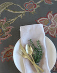Table setting with white plate, gold fork and spoon, and a sprig of thyme on a floral-patterned tablecloth.