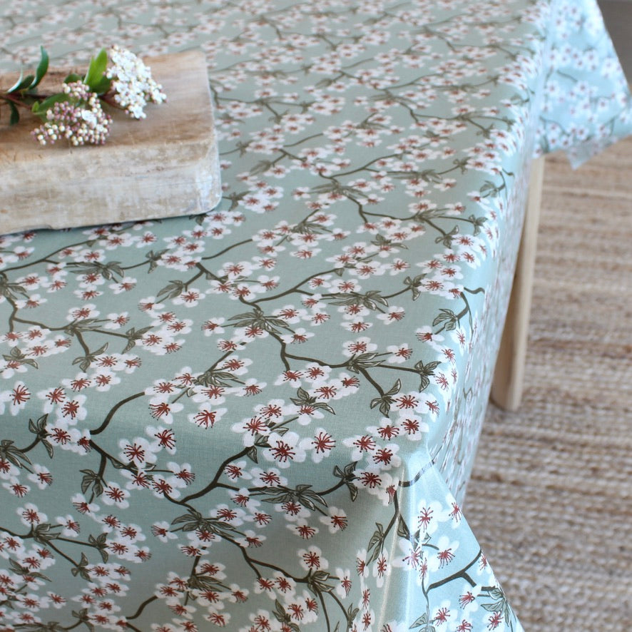 A table covered with a sage green tablecloth featuring a delicate white and pink cherry blossom branch pattern, styled with a wooden cutting board and white flowers.