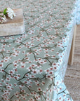 A table covered with a sage green tablecloth featuring a delicate white and pink cherry blossom branch pattern, styled with a wooden cutting board and white flowers.