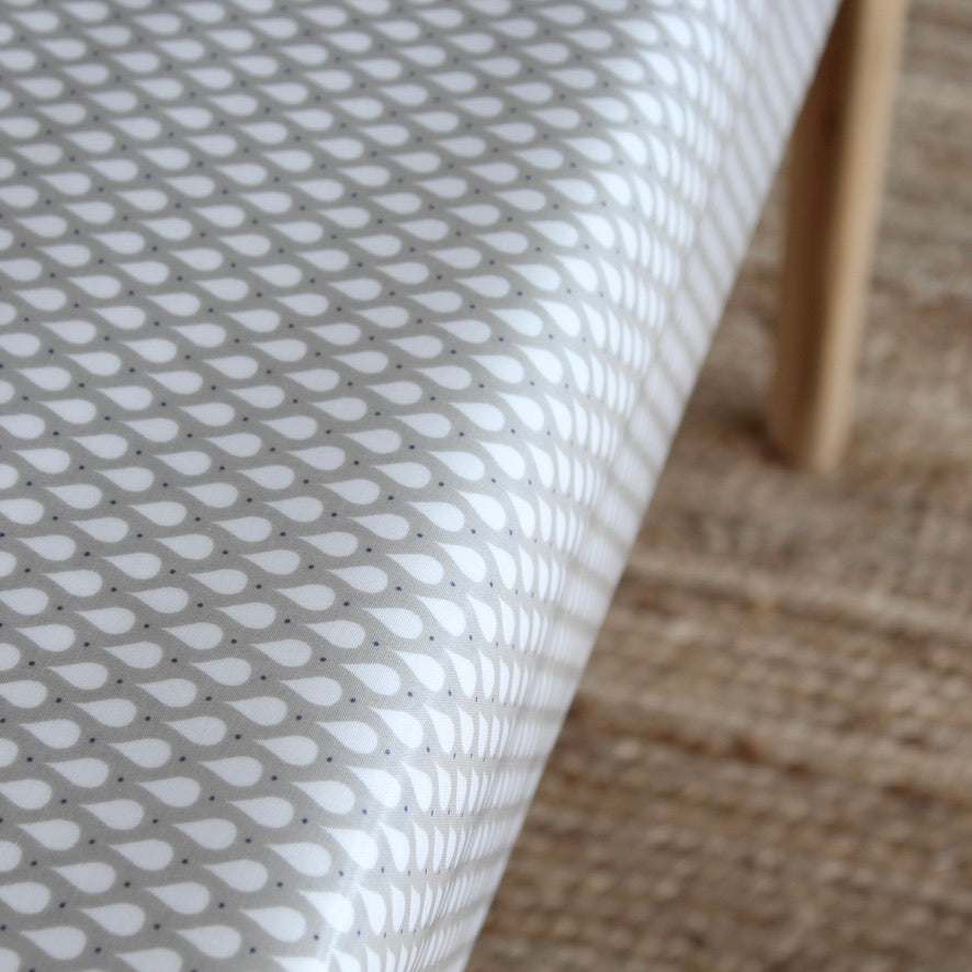 A close-up of a white tablecloth with a repeating gray teardrop pattern, draped over a table with a light wood leg visible on the right side.