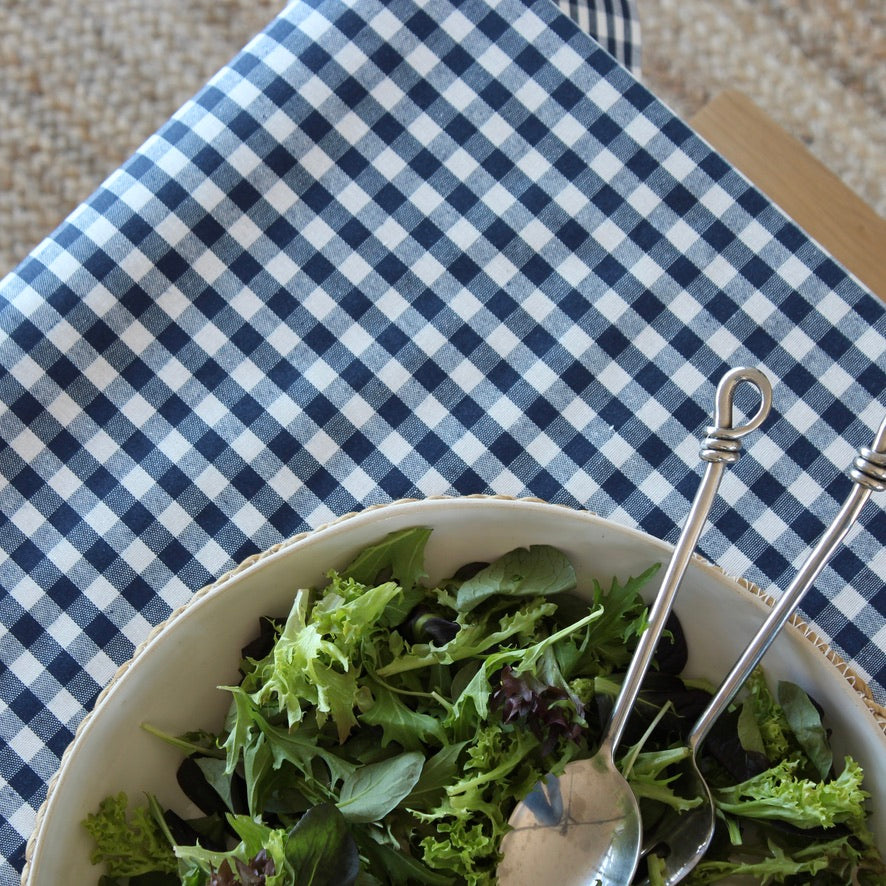 A close-up of a blue and white gingham checkered tablecloth with a white bowl of green salad and two metal serving spoons resting in it.