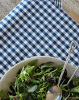 A close-up of a blue and white gingham checkered tablecloth with a white bowl of green salad and two metal serving spoons resting in it.