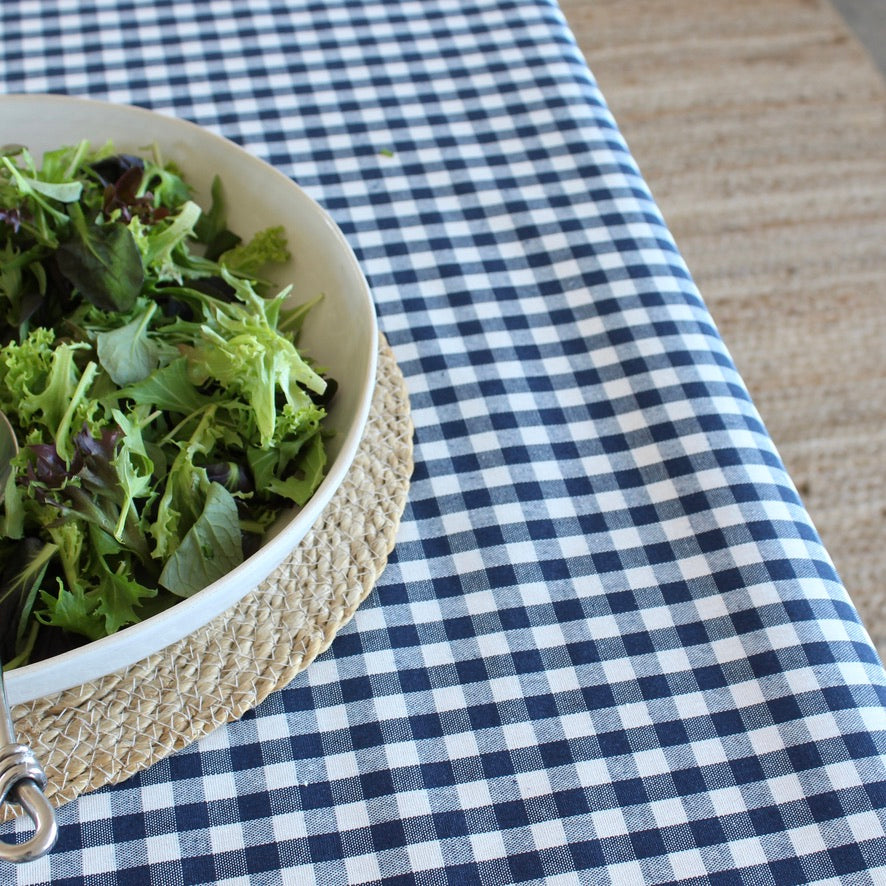 A navy and white gingham tablecloth spread across a wooden table, with a white bowl of mixed green salad resting on a woven beige placemat in the lower-left corner.