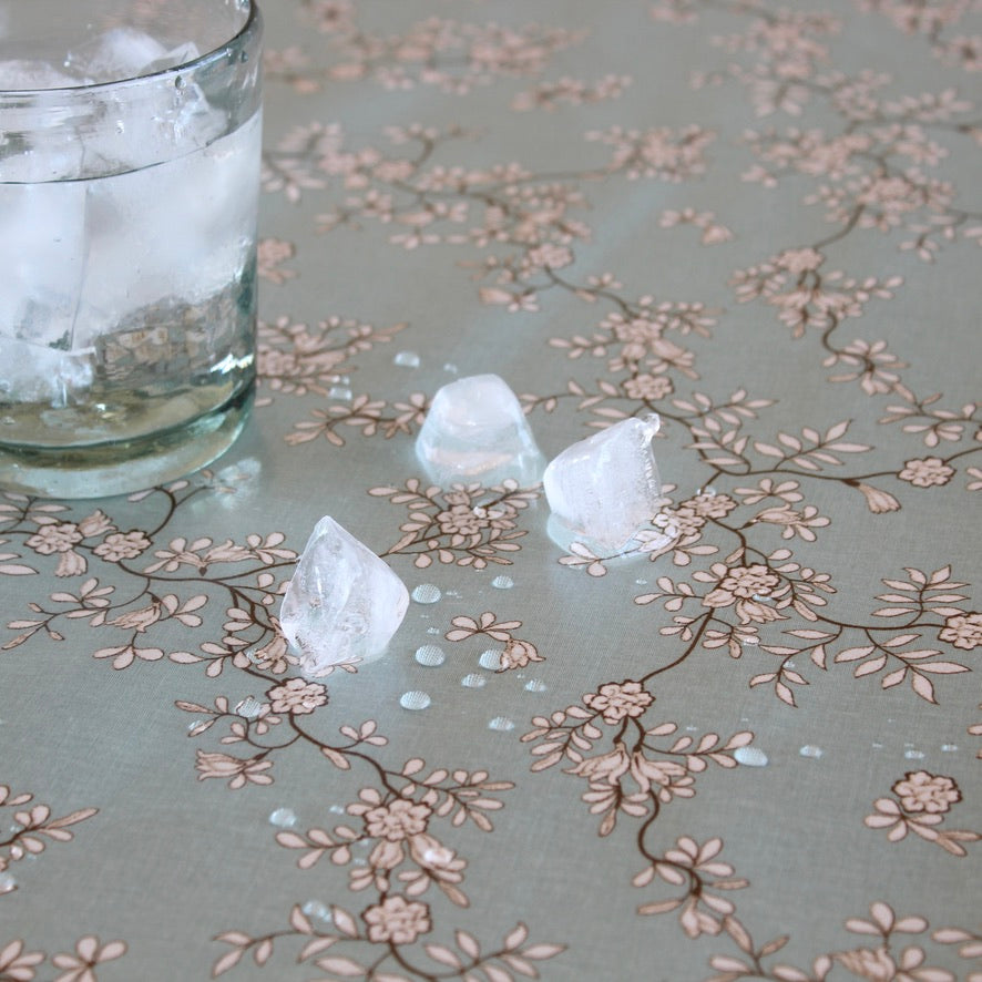 A close-up of a clear glass with ice on a light gray table covered in a delicate beige floral vine pattern, with three ice cubes scattered on the surface.