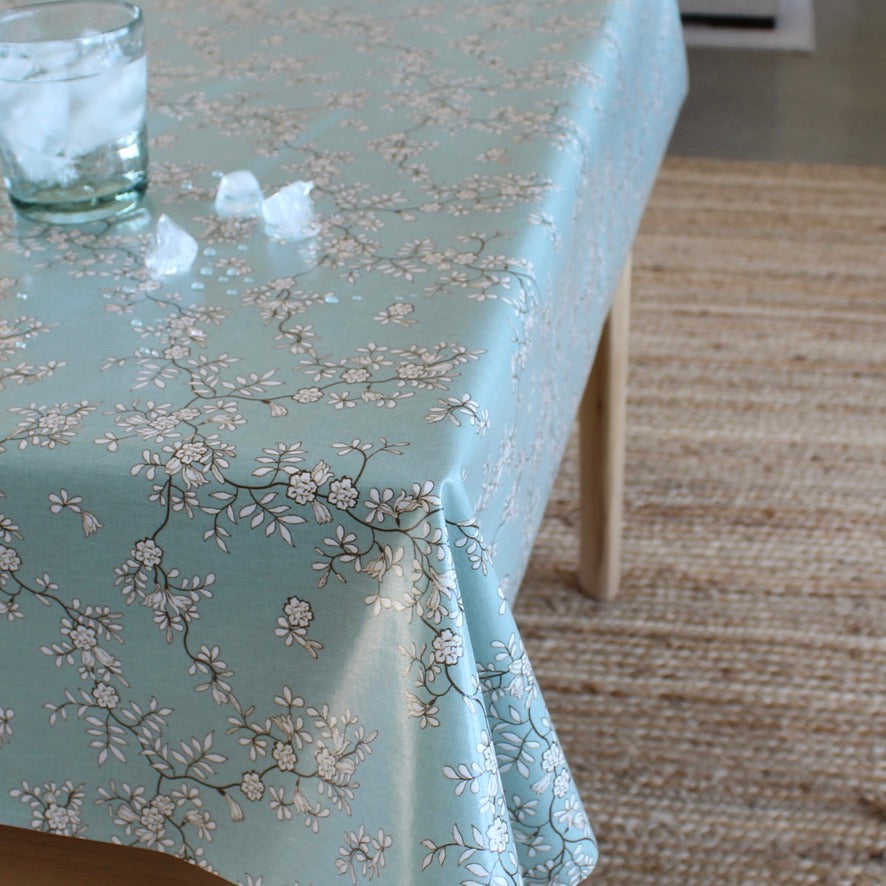 A light blue tablecloth with a delicate white and gray cherry blossom branch pattern, draped over a table in a home setting with a woven rug and a glass of water on top.