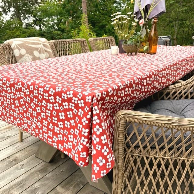 A red tablecloth with a white floral pattern draped over an outdoor wicker dining table on a wooden deck, surrounded by wicker chairs.